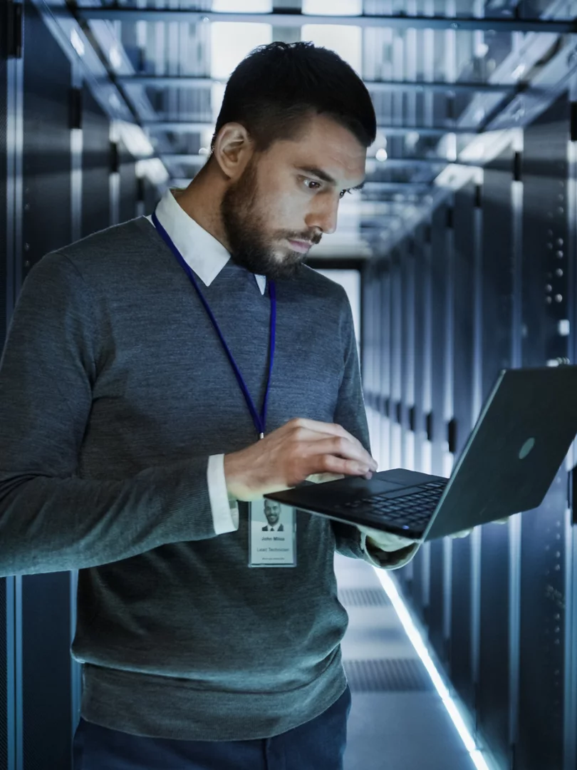 IT Technician Works on a Laptop in Big Data Center full of Rack Servers. He Runs Diagnostics and Maintenance, Sets up System.